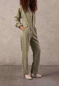 Osoba ubrana w oliwkowy kombinezon z guzikami, z podwiniętymi rękawami, oraz beżowe, wysokie botki za kostkę, stojąca przy neutralnej ścianie.