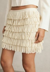 Mini gonna con frange in crema, con strati di anelli di tessuto che creano un aspetto testurizzato. Vita elastica per comfort e vestibilità.
