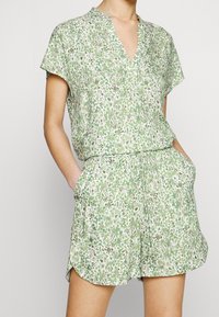 Grembiule corto con motivo verde e bianco, maniche corte, scollo a V, vita elastica e tasche laterali. Tessuto leggero con un design giocoso.