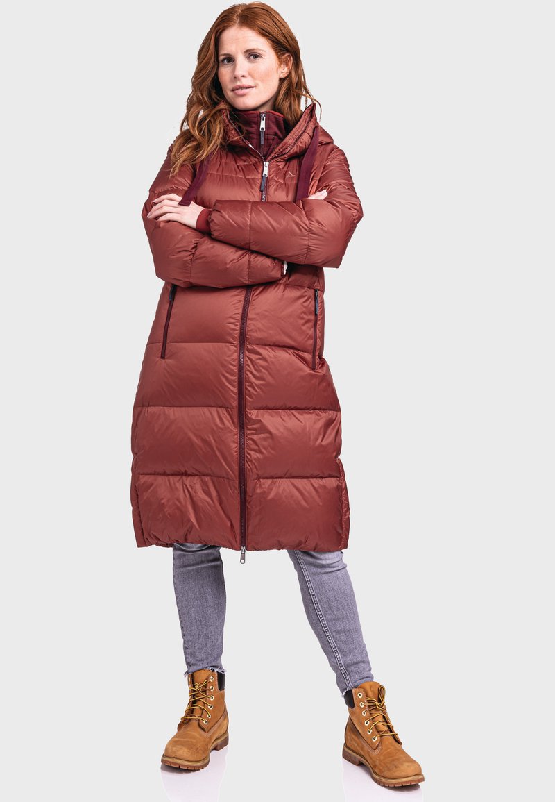 Roodharige vrouw in een lange bordeaux pufferjas, grijze jeans en tan-gekleurde veterschoenen, staande met de armen over elkaar tegen een lichte achtergrond.