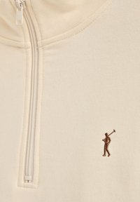 Sudadera con cremallera color beige fabricada en tejido suave, que presenta una pequeña figura marrón bordada sosteniendo un palo en el área del pecho izquierdo.