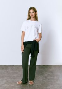 Camiseta blanca de manga corta con la palabra "BLOUNIE" en letras metálicas, combinada con pantalones de pierna ancha de color verde oscuro y un suéter negro sobre la cintura.