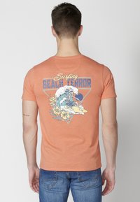 Narančasta pamučna majica s grafikom koja prikazuje kostura kako surfuje unutar trokuta, okruženog cvijećem i natpisom "Surfing Beach Terror."