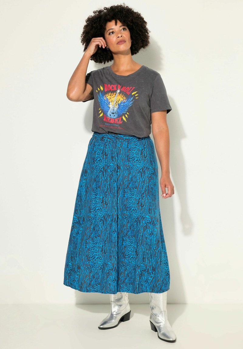 T-shirt graphique en charbon avec un imprimé vibrant, associé à une longue jupe bleu et noire à motifs et des bottes de cowboy argentées brillantes.