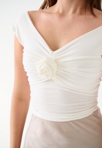 Top bianco con maniche a cappuccio, caratterizzato da un design arricciato e un dettaglio centrale a forma di rosa in tessuto. Tessuto liscio, abbinato a una gonna beige.