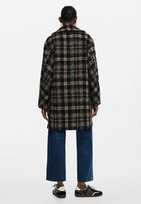 Long, oversized black and multicolored checkered coat with a collar, textured fabric, and dropped shoulders, paired with wide-leg jeans and sneakers.