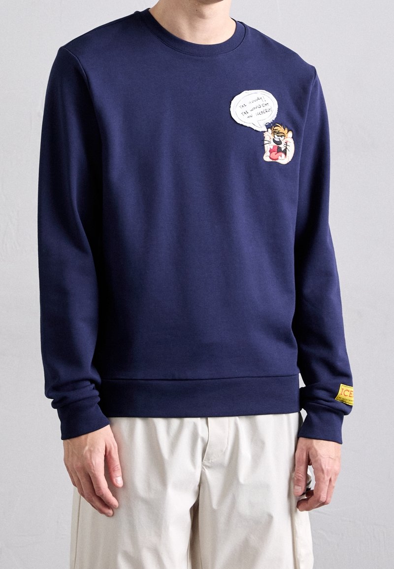 Sweat-shirt bleu marine avec patch tigre de dessin animé et bulle de dialogue sur la poitrine, patch jaune au poignet gauche, porté avec un pantalon beige clair.