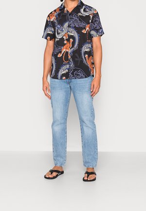 Zwart shirt met korte mouwen en levendige oranje en blauwe draak- en tijgerpatronen, gecombineerd met lichtblauwe jeans en zwarte slippers.