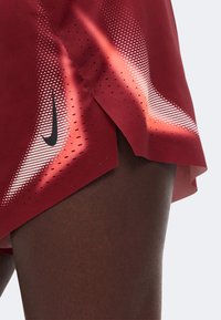 Κοντινό πλάνο στο μηρό ενός ατόμου που φοράει μαρόν αθλητικό σορτς με μαύρες τρύπες και ένα λευκό μοτίβο με διαβάθμιση που φέρει το μαύρο λογότυπο του Nike.