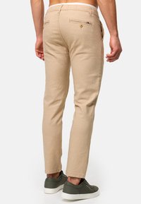 Beige bukser laget av et teksturert stoff, med baklommer med knapper, slim fit og oppbrettede nederkantar. Båret med mørkegrønne sneakers.