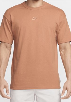 Homme portant un t-shirt Nike marron clair uni à manches courtes et un short blanc, vu du cou jusqu'aux hanches, les bras le long du corps.