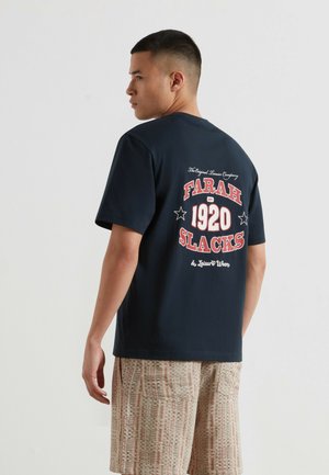Mężczyzna zwrócony bokiem, ubrany w granatowy t-shirt z dużym czerwono-białym napisem „Farah 1920 Slacks” i wzorzyste beżowe szorty.