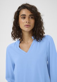 Haut bleu clair à manches longues avec col en V, présentant une texture lisse et un plis subtils aux épaules. Design simple et minimaliste.