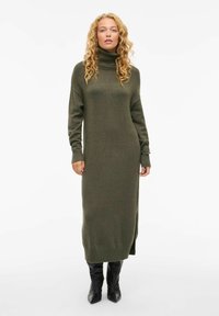 Robe en tricot côtelé vert olive avec col roulé, à manches longues et fentes latérales, atteignant sous le genou. Assortie à des bottes noires montantes.