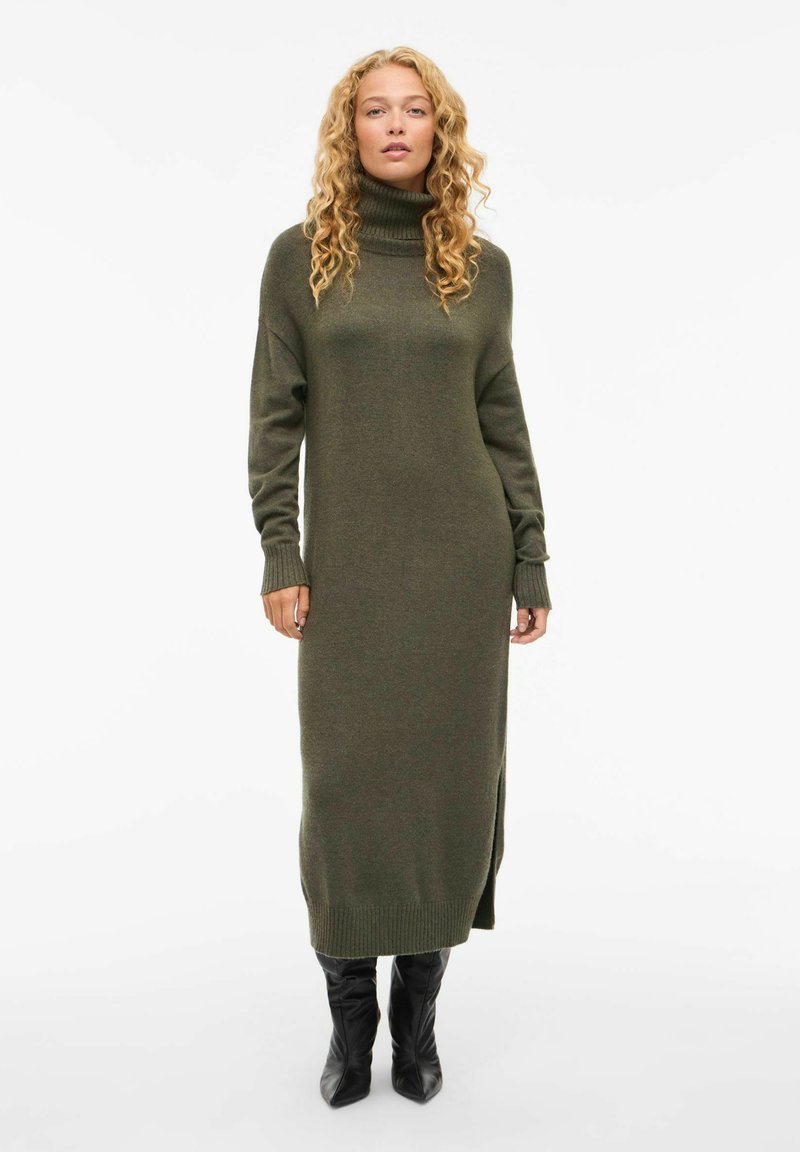 Olive roheline, sooniknito turtleneck kleit pikkade varrukatega ja külgslitidega, ulatudes üle põlve. Kombineeritud mustade põlve kõrguste saapadega.
