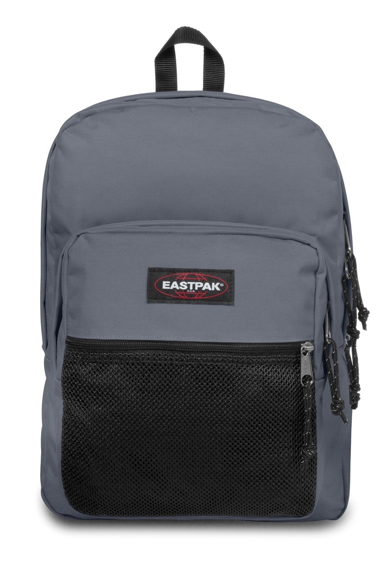 Mochila de tela gris con un bolsillo frontal de malla, asa superior y cremalleras. Presenta un parche de logo negro y correas ajustables.