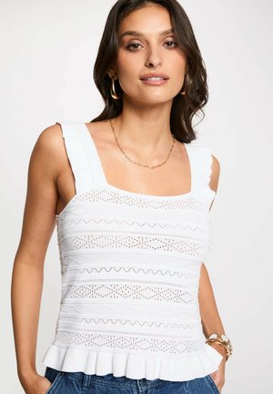 Donna che indossa un top bianco lavorato a maglia senza maniche con ritagli decorati, orlo arricciato, orecchini a cerchio dorati, collana con catena e braccialetti su uno sfondo semplice.