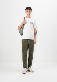 Giovane uomo con occhiali che indossa una t-shirt bianca con logo Pepsi Cola, pantaloni verde oliva, sneakers bianche e porta una borsa a tracolla grigia su sfondo bianco.