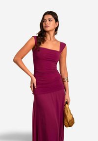 Figurbetontes burgunderfarbenes Kleid mit quadratischem Ausschnitt und kurzen Ärmeln, das eine geraffte Textur aufweist. Das Model hält eine goldene Clutch.