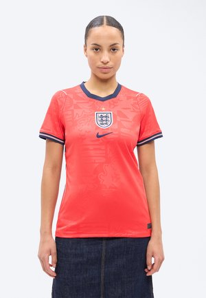 Femeie privindu-și înainte, purtând un tricou de fotbal roșu al Angliei cu margini bleumarin și o fustă din denim închis, pe un fundal simplu și deschis.