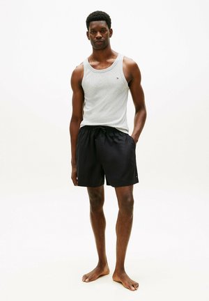 Man staat op blote voeten, draagt een lichtgrijs shirt zonder mouwen en zwarte short met trekkoord, tegen een effen witte achtergrond.