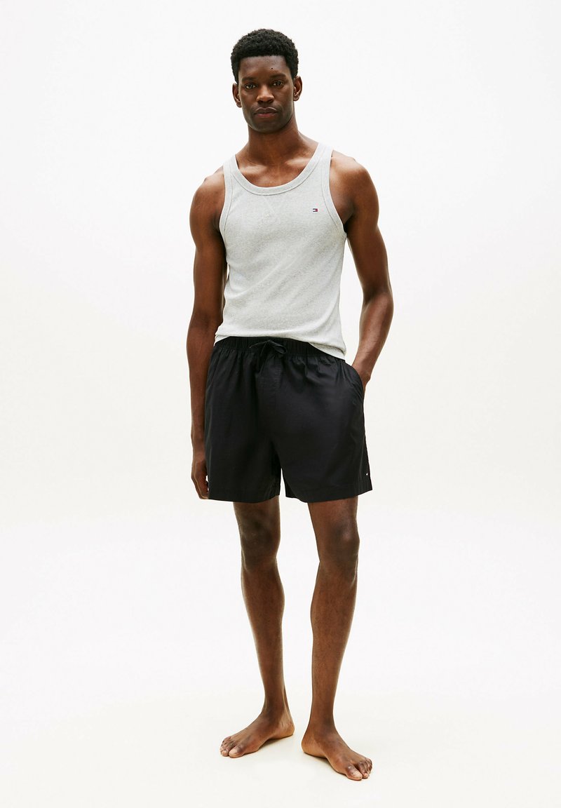 Mężczyzna stojący boso, ubrany w jasnoszary top na ramiączkach i czarne szorty ze sznurkiem na gładkim białym tle.