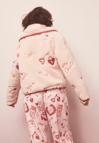 Roza puhasta jakna z rdečim srcem in vezeninami na temo ljubezni, v kombinaciji z ujemajočimi hlačami za pižamo z igrivim, vzorčastim dizajnom.