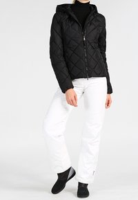 Veste matelassée noire avec capuche, fermeture éclair frontale et poches latérales, accompagnée d'un pantalon de neige blanc et de bottines noires.