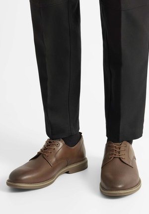 Brązowe skórzane buty o gładkim wykończeniu, zaokrąglonym nosku, fakturowanej cholewce i sznurowanym fasonie; w połączeniu z czarnymi spodniami i ciemnymi skarpetkami.