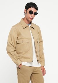 Man wearing beige jacket and pants, white shirt, and black oval sunglasses, standing with one hand in his pocket against white background.