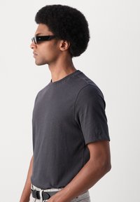 T-shirt gris à manches courtes en tissu doux, avec un col rond et une coupe décontractée, associé à des lunettes de soleil noires et un jean de couleur claire.