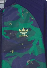 Adidas-jacka med en grön och blå camouflage-design och en slät textur. Guld-logotypdetaljer nära dragkedjan. Tillverkad av lättviktsmaterial.