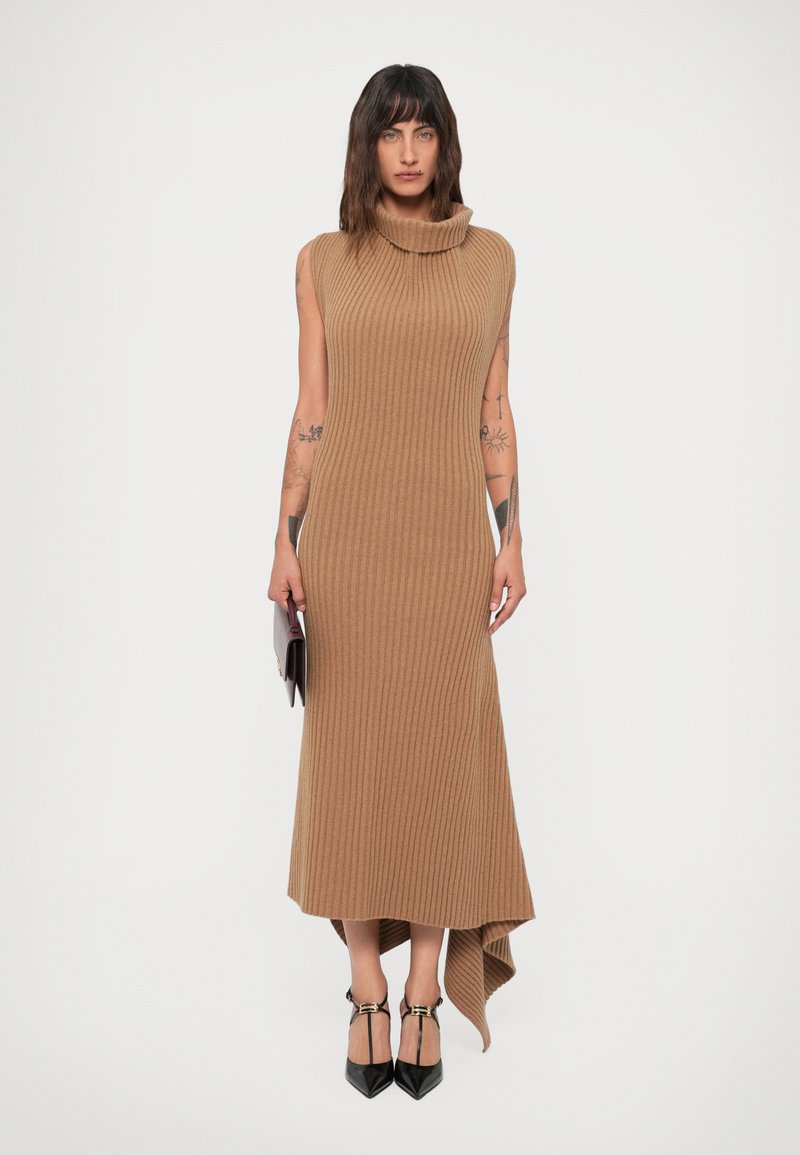 Femme avec des tatouages portant une robe midi sans manches en col roulé côtelé de couleur camel, tenant une pochette et portant des escarpins noirs à talons pointus.