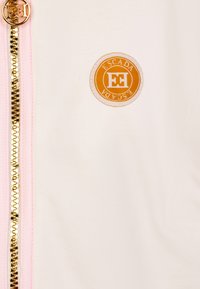 Giacca bianca a zip con zip dorato, caratterizzata da un logo rotondo arancione ESCADA sul lato sinistro e dettagli rosa sui bordi della zip.