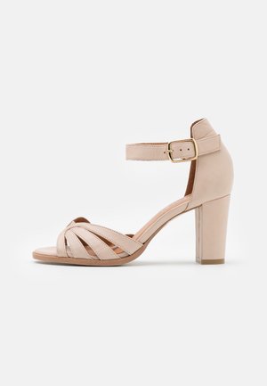 Beżowy sandał na obcasie z paskiem wokół kostki, z krzyżującym się wzorem na górze, zamkniętą piętą i masywnym, blokowym obcasem. Gładka skóra.