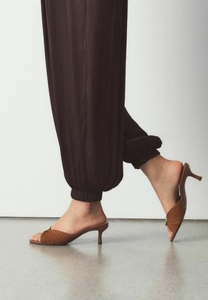 Person iført brune, samlet ankelbukser og brune kitten heel-sandaler med åben hæl, stående på en glat gulvbelægning.