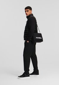 Vesta puffer neagră peste un tricou cu mânecă lungă negru, combinată cu pantaloni negri și teniși negri cu talpă albă. Geantă de umăr neagră expusă.