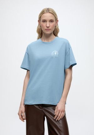 Vrouw met lang blond haar, gekleed in een lichtblauw T-shirt met wit logo en donkerbruine leren broek, staande tegen een effen witte achtergrond.