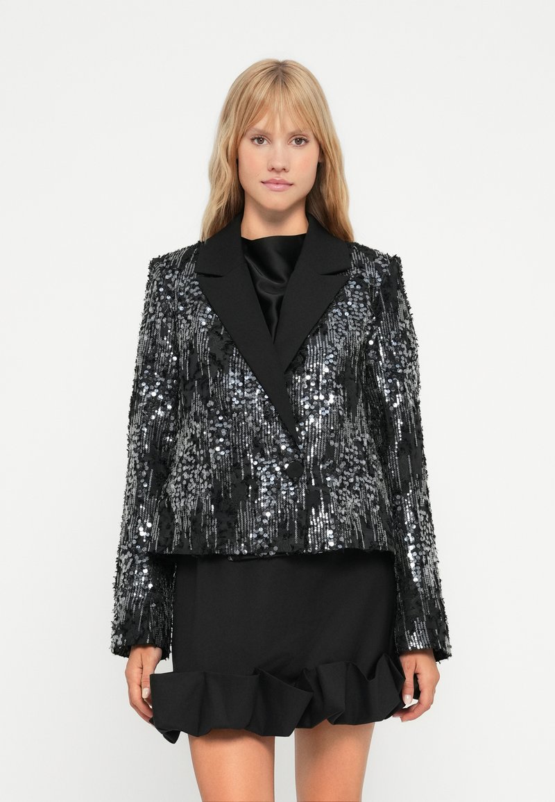 Zwarte blazer met pailletten, glanzende textuur, scherpe revers en een cropped ontwerp. Gecombineerd met een zwarte rokje met ruches.