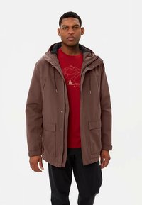 Bruine waterdichte jas met capuchon, met zakken aan de voorkant en trekkoorden, gedragen over een rood T-shirt met grafische opdruk. Effen achtergrond.