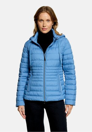 Femme portant une veste matelassée bleu clair avec capuche, un col roulé noir et un pantalon foncé, debout devant un fond clair uni.
