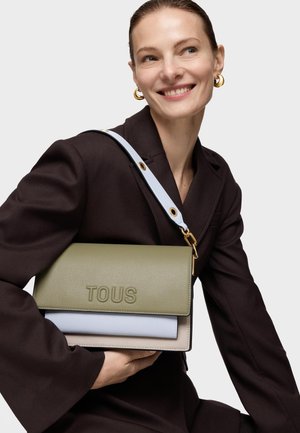 Mujer con blazer oscuro sonriendo, sosteniendo un bolso tricolor de TOUS con paneles en oliva, azul claro y beige, y una correa blanca sobre el hombro.