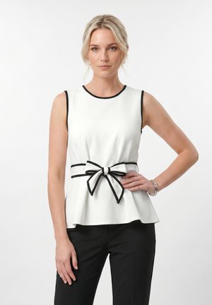 Plavokosa žena stoji s rukom na kukovima, nosi bijelu bluzu bez rukava s crnom obrubom i mašnom u struku, uparenu s crnim hlačama.