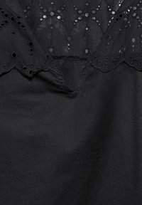 Blouse noire avec un col en forme de vagues, ornée de motifs brodés en œillets et de détails découpés, fabriquée en tissu léger.