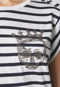 Camiseta a rayas con rayas horizontales en gris y azul marino. Adornada con un diseño de calavera metálica con cuentas en el pecho, añadiendo textura.