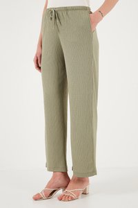 Naine kannab heleoliivirohelisi tekstuuriga paeltega pükse, mille sääretel on lõhikud, ja beeže avatud varbaga kontsakingi, seisab ühe käega taskus.