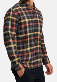 Homme portant une chemise à manches longues boutonnée avec un motif à carreaux noir, rouge, jaune et gris, les mains partiellement dans les poches d’un pantalon noir.