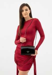 Černá lakovaná kabelka s řetízkem, geometrického tvaru, s embosovaným logem včely. Nosičena s přiléhavými červenými šaty.