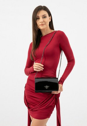 Schwarze lackierte Lederhandtasche mit einer Kettenriemen, geometrischer Form und eingeprägtem Bienensymbol. Getragen zu einem figurbetonten roten Kleid.