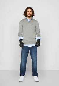 Grijze trui met rits over een lichtblauw overhemd, gecombineerd met een blauwe spijkerbroek en witte sneakers. Het model draagt zwarte handschoenen.